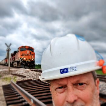 Photographer wearing construction hard hat near railroad crossing in rural area | P-Tn